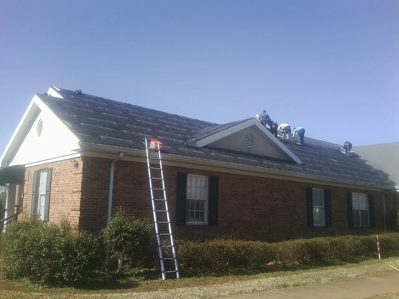 Crew tearing shingles off a brick ranch roof with a ladder set — Emergency Roof Repair work by our crew in Kings Park