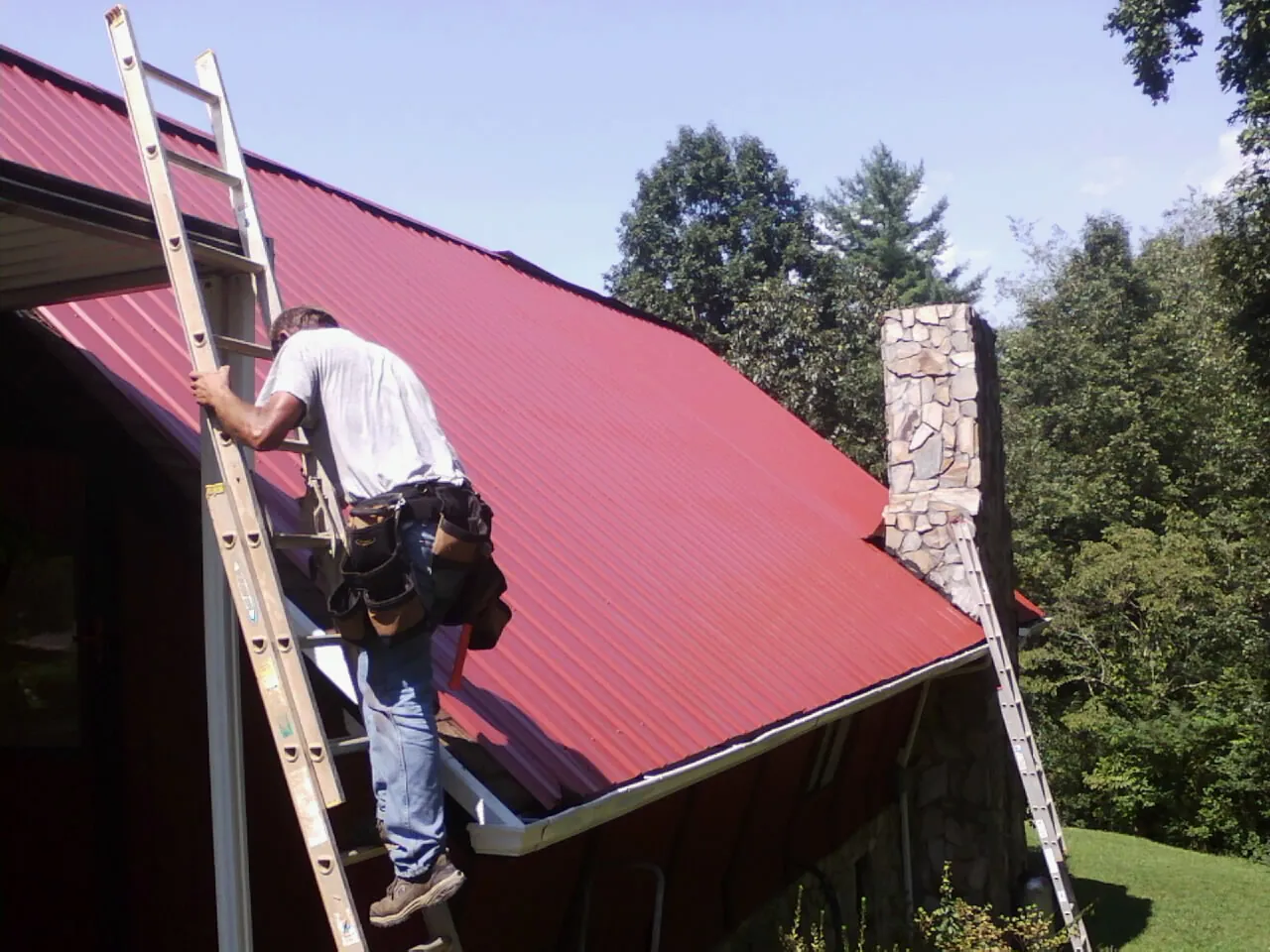 Roofer on a ladder beside a red metal roof and stone chimney — Soffit & Fascia Repair by Roofing Brothers in Kings Park